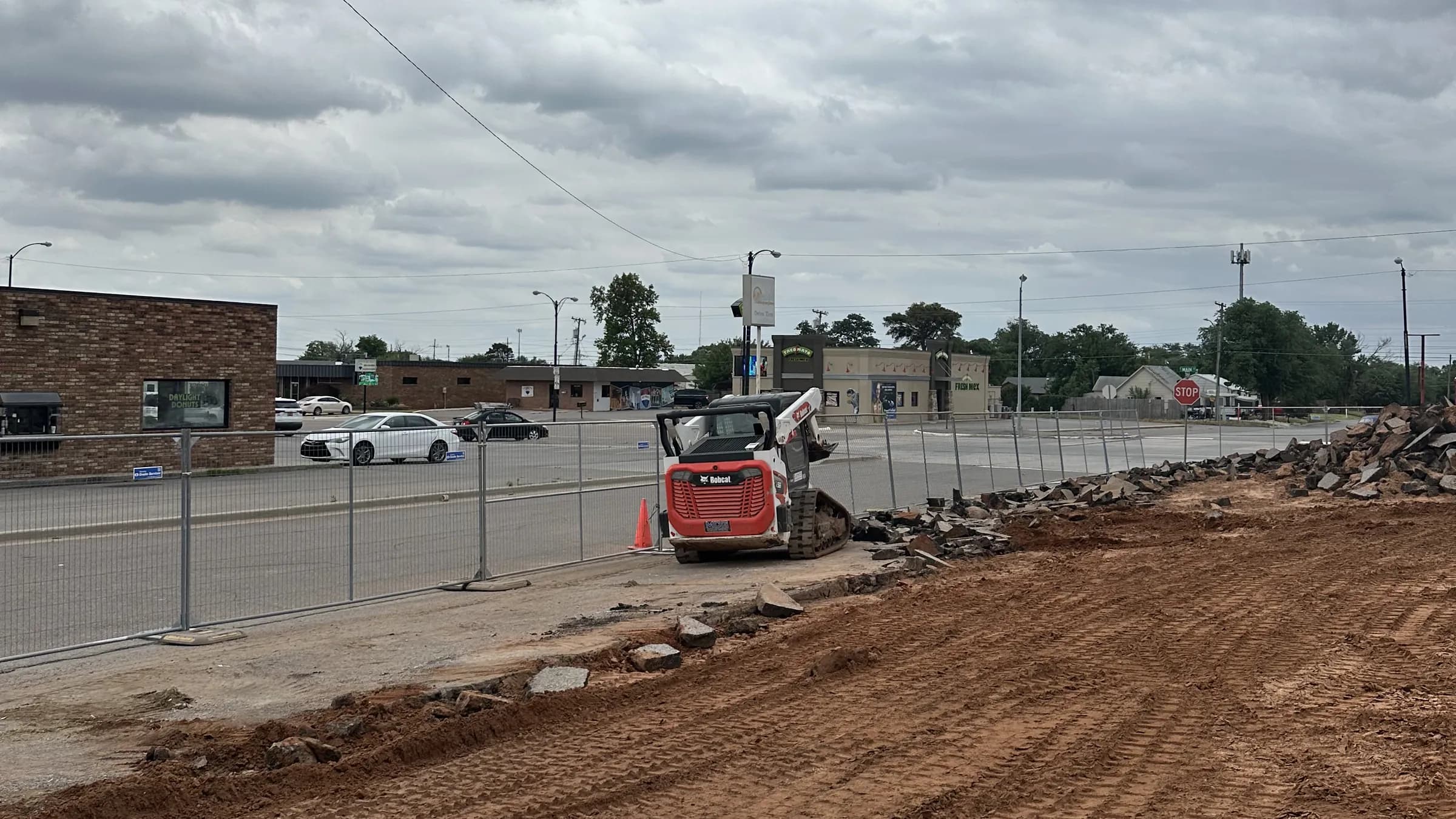 A compact track loader spreading red-dirt fill across a fenced commercial lot where a foundation was removed, the brick storefronts of the main street behind it under a heavy overcast sky.