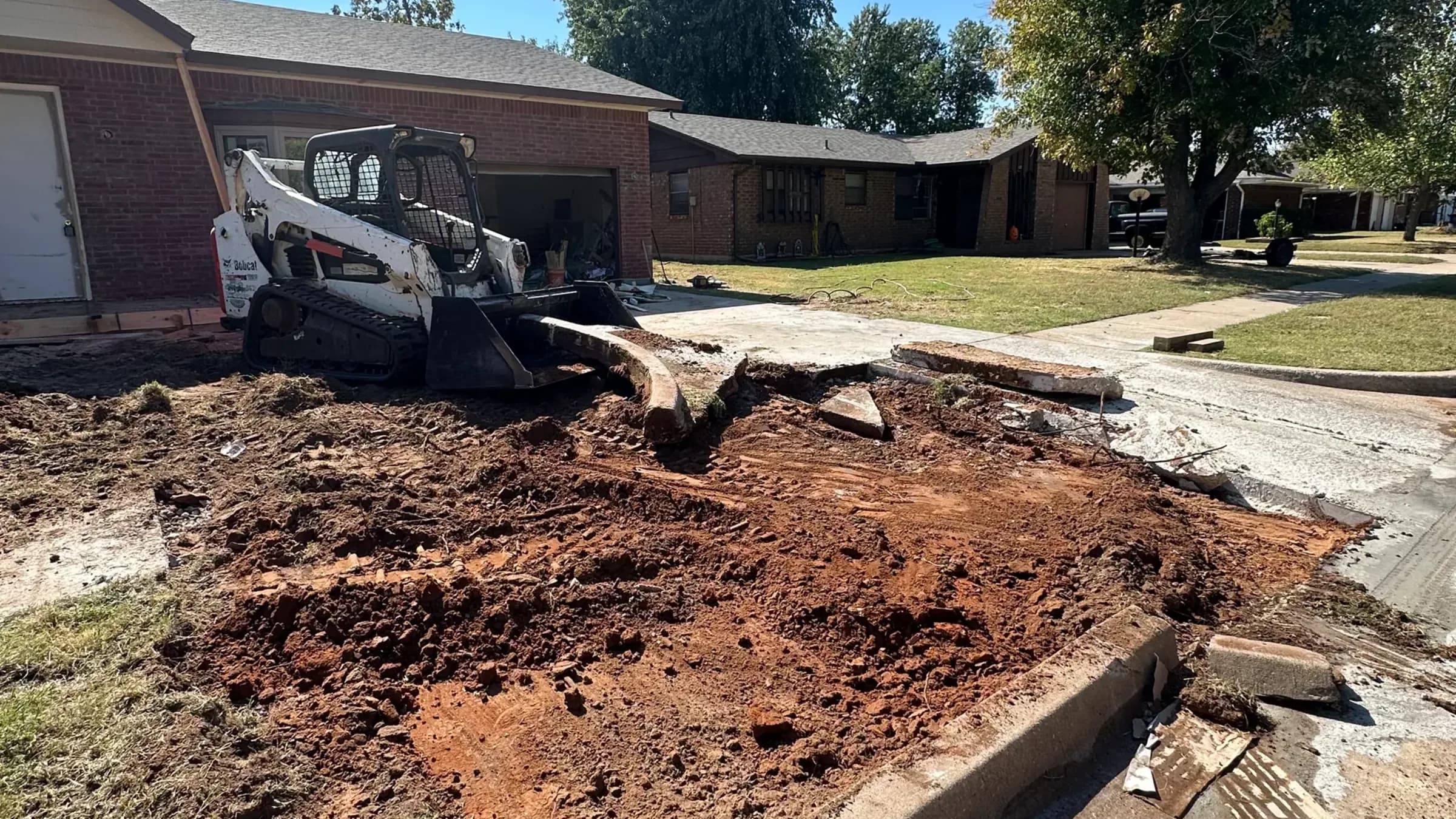 A compact track loader breaking a concrete driveway out at the curb line, broken slab sections and red dirt where the drive used to run.