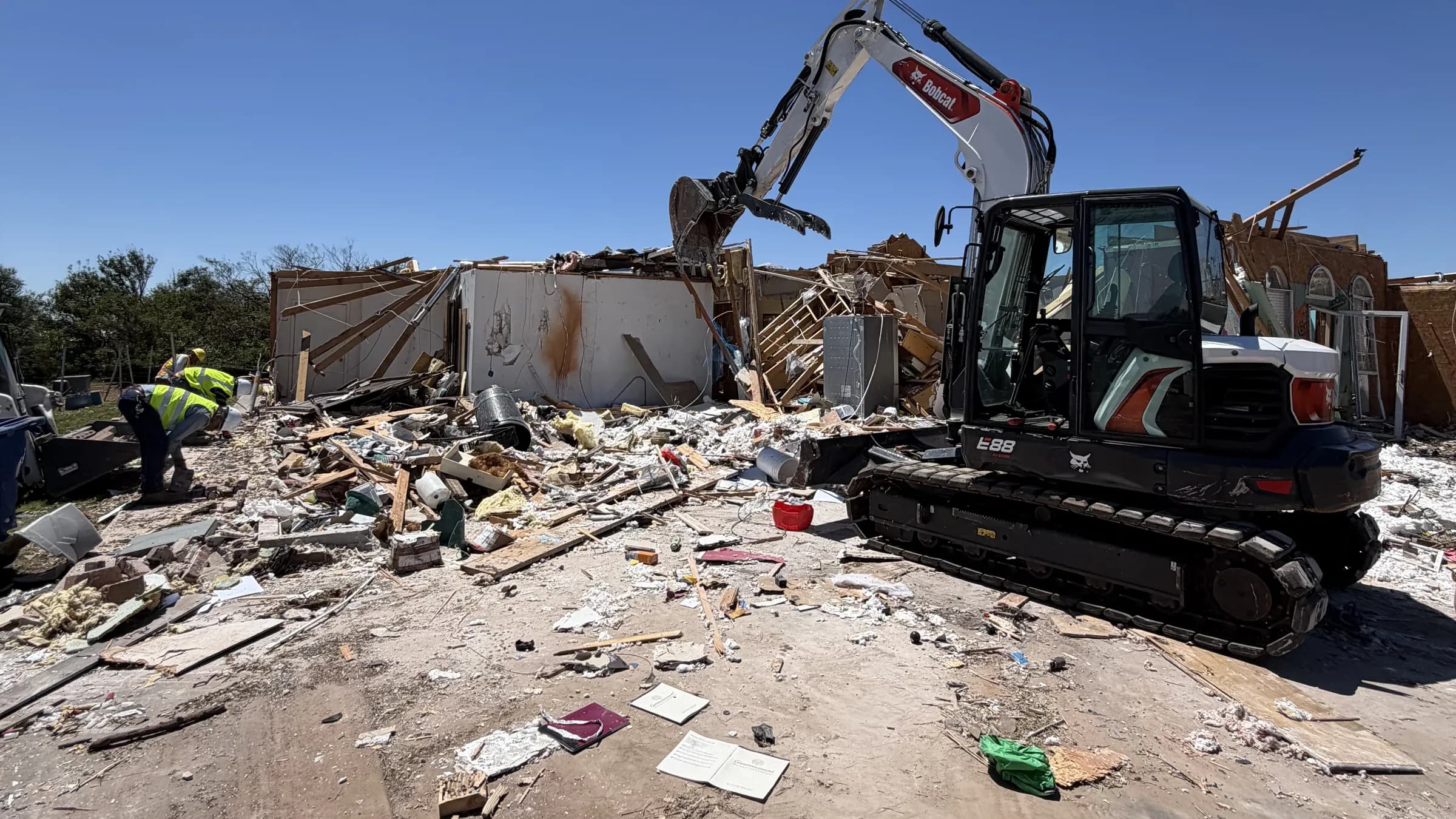Una excavadora compacta tumbando el último muro en pie de un edificio arrasado, con el escombro regado por el lote y un miembro del equipo en chaleco reflejante a la orilla