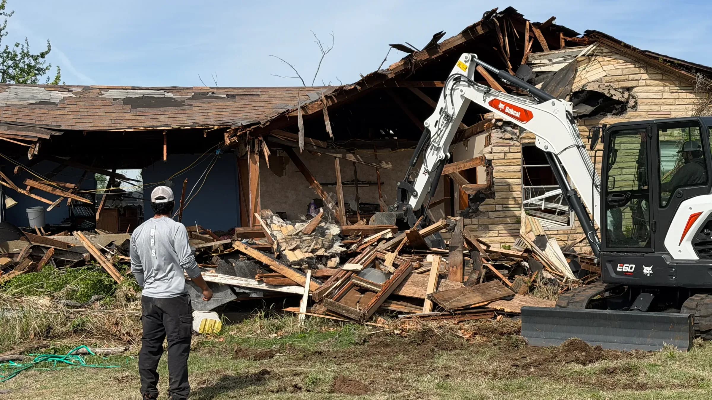 A compact excavator pulling the roof off a stone-faced ranch house, timbers and roofing collapsed into the yard while a crew member watches from the grass.