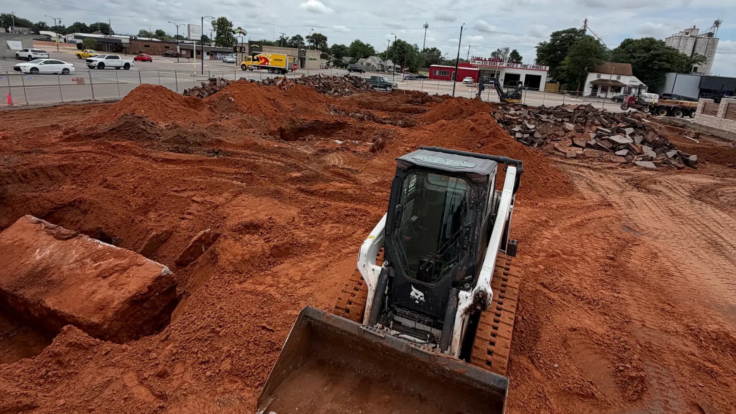 Un minicargador de orugas con bote de dientes trabajando un lote comercial ya limpio, con montones de arcilla colorada y concreto quebrado junto a la cerca del fondo