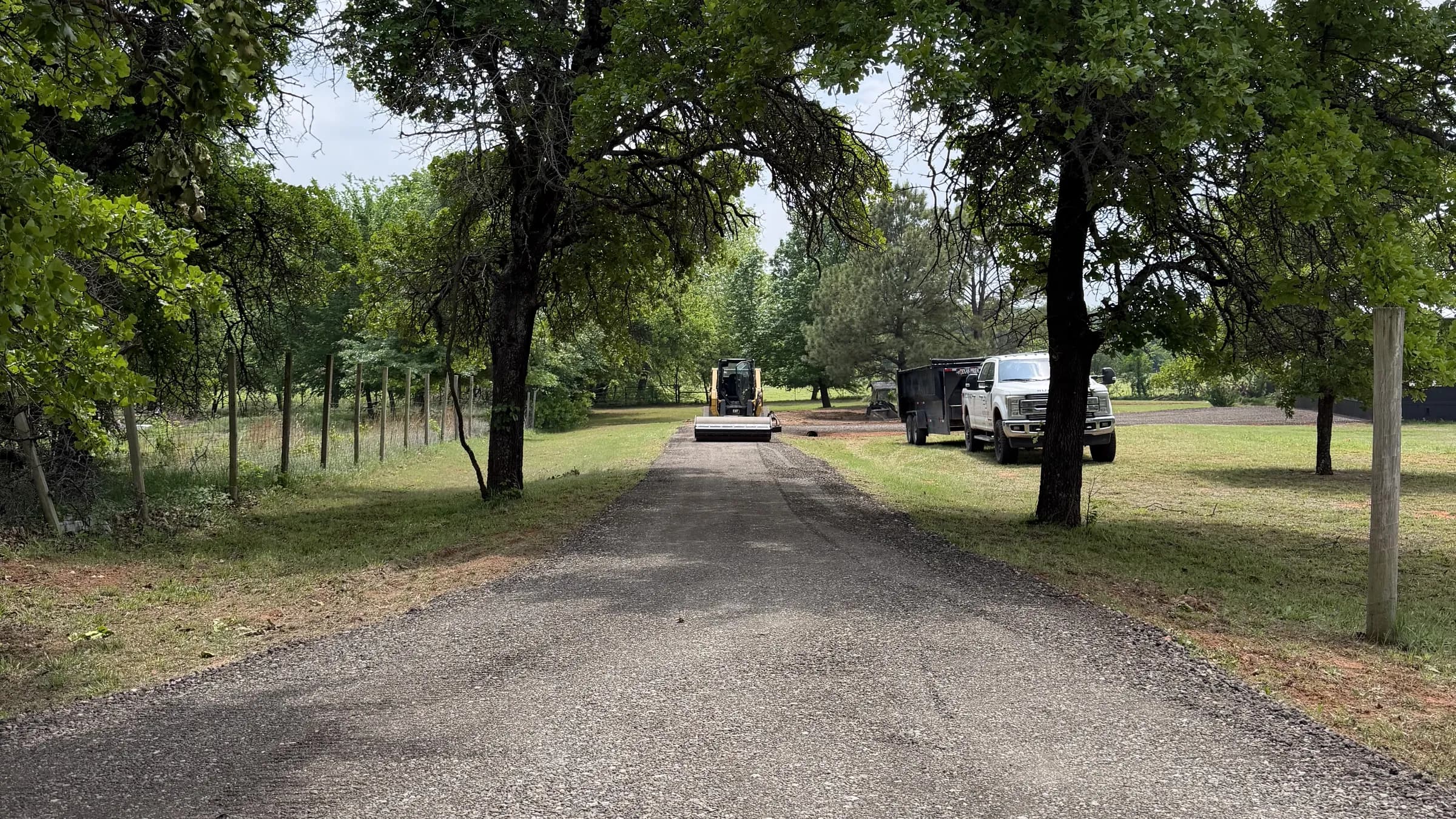 A tree-lined driveway freshly resurfaced with dark asphalt millings, a roller and dump trailer working at the far end