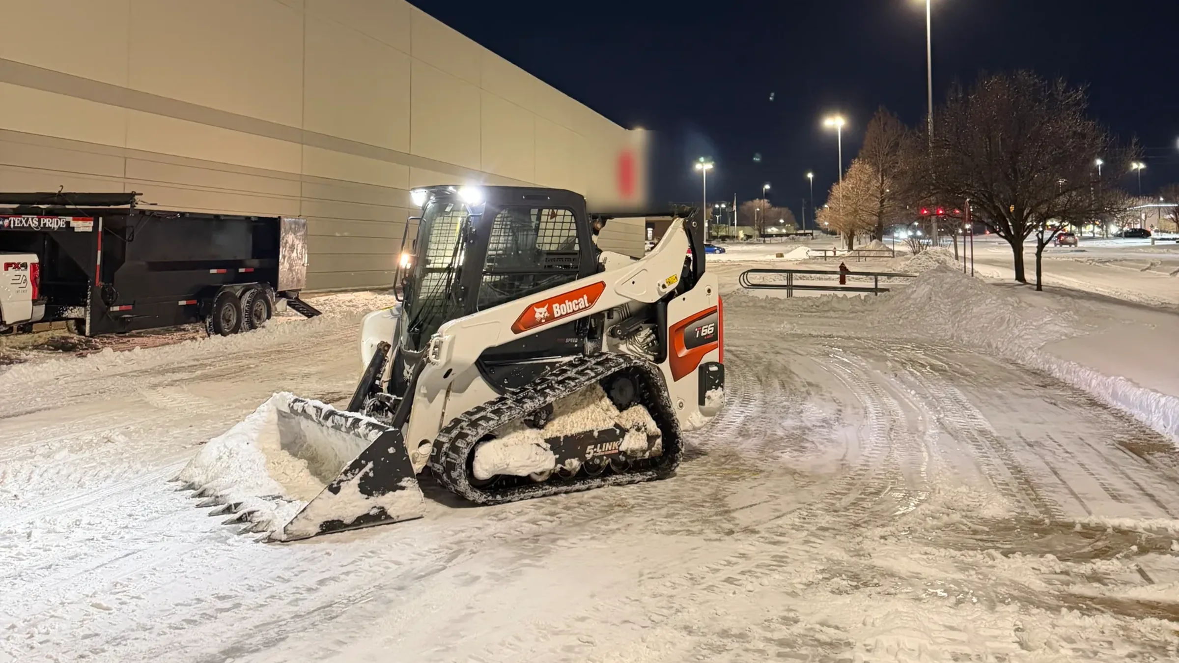 Un minicargador de orugas empujando nieve en un estacionamiento comercial iluminado ya de noche, con los carriles despejados y la nieve amontonada en las orillas